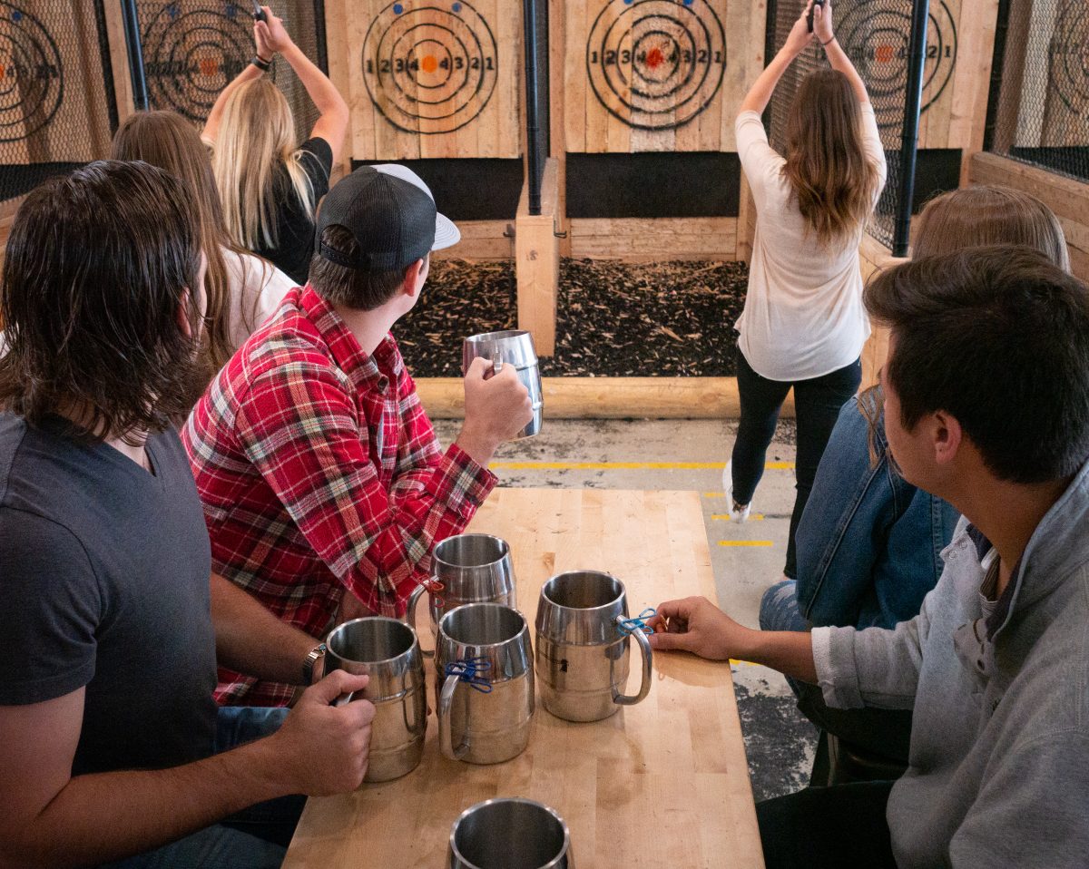 a group of people drinking soda from metal mugs while two women throw axes at targets.
